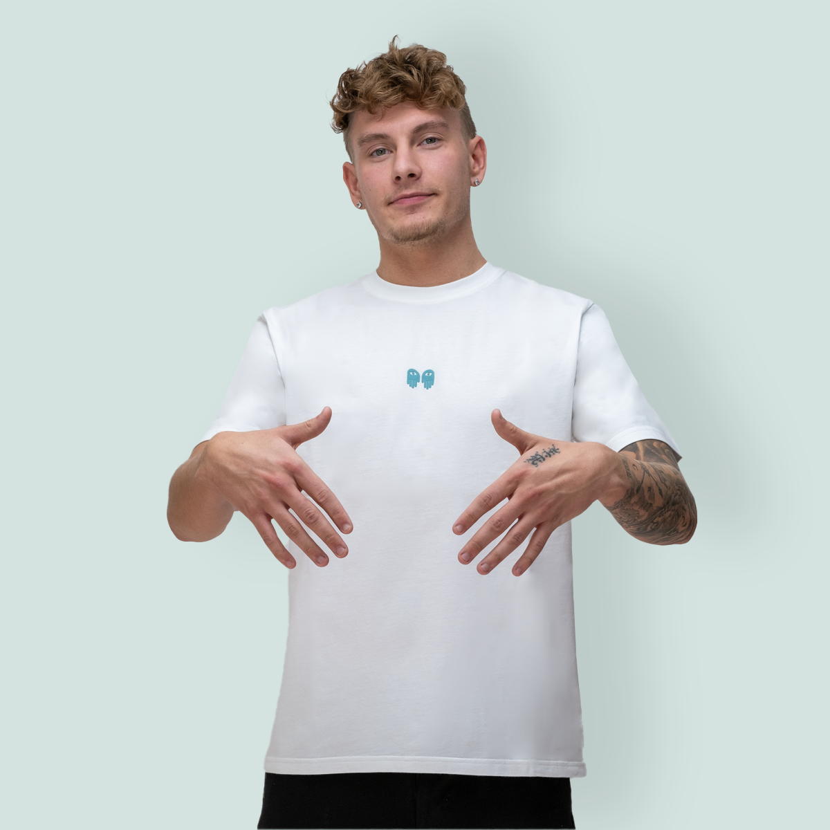 En ung mann iført en t-skjorte med et logo i midten av brystet, poserer med begge hendene oppe foran en lysgrønn bakgrunn.
